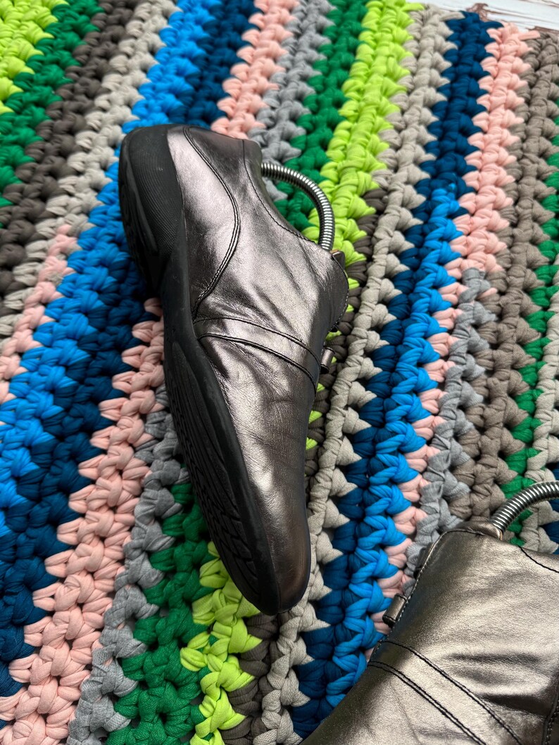 May include: A pair of silver leather shoes with a zipper on the side. The shoes are on a colorful crocheted rug.