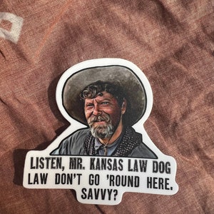 May include: A white sticker with a cartoon illustration of a man wearing a cowboy hat and a beard. The text on the sticker reads "LISTEN, MR. KANSAS LAW DOG LAW DON'T GO 'ROUND HERE. SAVVY?"