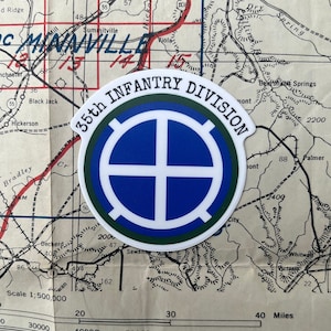 May include: A circular sticker featuring the emblem of the 35th Infantry Division. The design includes a blue and white cross within a blue circle, surrounded by a green border. The words "35th INFANTRY DIVISION" are arched around the top edge. The sticker rests on a vintage map.