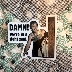 May include: A sticker featuring a cartoon image of a man peeking around a wooden post. The sticker has the text "DAMN! We're in a tight spot." The sticker is on a patterned fabric background.