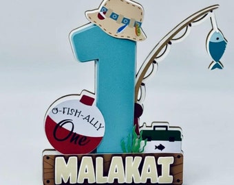 Puede incluir: Adorno para tarta de primer cumpleaños con temática de pesca. El número uno es azul claro, con un sombrero de pescador encima. Una caña de pescar con un pez azul cuelga del número. El nombre "MALAKAI" está en la base.