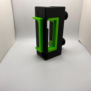 May include: A black rectangular phone mount featuring bright green adjustable brackets on the front and two black circular adjustment knobs on the side. The text IBP is printed in black on the upper left corner of the main black housing structure.