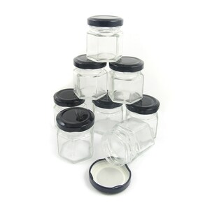 May include: A stack of ten clear glass hexagonal jars with black lids. The jars are arranged in a pyramid shape, with the top jar being the smallest and the bottom jars being the largest. One lid is laying on its side.