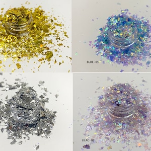 May include: Four small jars of iridescent glitter flakes in gold, blue, silver, and lilac. The glitter is scattered around the jars.