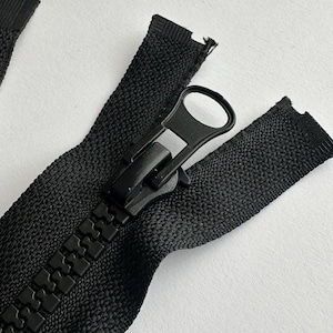 May include: Black zipper with a black plastic slider and pull tab. The zipper is closed and the slider is in the middle of the zipper.