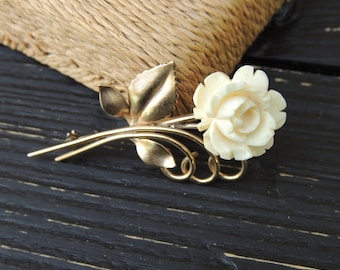 May include: A gold tone brooch featuring a carved white rose with leaves and a stem.