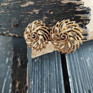 Op de afbeelding: Een paar gouden clip-on oorbellen met een bloemmotief. De oorbellen zijn gemaakt van metaal en hebben een swirl in het midden.