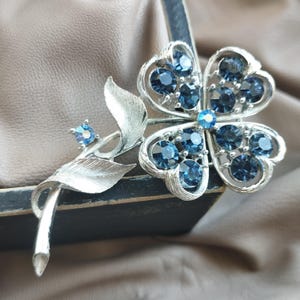 Puede incluir: Broche plateado con diseño floral, con forma de trébol de cuatro hojas adornado con piedras preciosas azul oscuro. El broche incluye un tallo con hojas y un pequeño acento de piedras preciosas. El broche está sobre un fondo neutro.