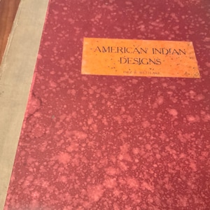 May include: A red book with a title that reads "American Indian Designs" in orange lettering. The book cover has a speckled pattern.
