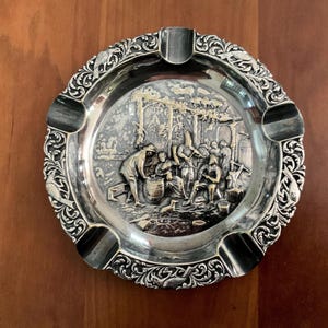 May include: A round silver-toned metal ashtray featuring a central embossed scene of figures gathered around a barrel under a trellis. The wide, ornate border is decorated with scrolling foliage and bird motifs, with four evenly spaced notches.