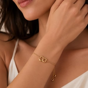 May include: A gold-toned chain bracelet featuring a central letter B charm with a textured surface. The bracelet includes a delicate link chain and an adjustable sliding mechanism with two small dangling heart-shaped charms at the ends.