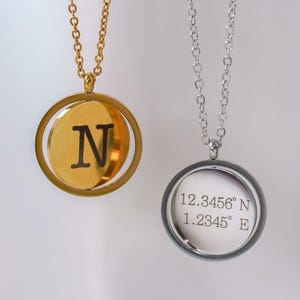 May include: Two pendant necklaces. One is gold-toned with the letter "N" on a circular pendant. The other is silver-toned with the coordinates "12.3456° N 1.2345° E" printed on a circular pendant. Both necklaces hang from delicate chains.