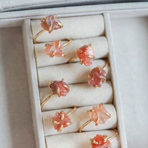 May include: A collection of seven gold-toned rings, each featuring a single raw, unpolished orange-red gemstone held in a prong setting. The rings are arranged in a series of padded slots, showcasing the natural, irregular texture of the stones.