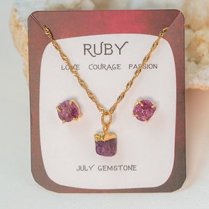 May include: A gold necklace and earrings set with a ruby pendant and earrings. The card reads "Ruby" and "Love, Courage, Passion" and "July Gemstone".