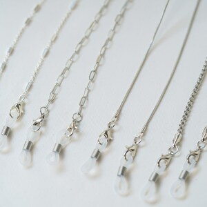May include: Seven silver eyeglass chains with clear plastic clasps. The chains are different styles, including a thin chain, a thicker chain, and a chain with small links.