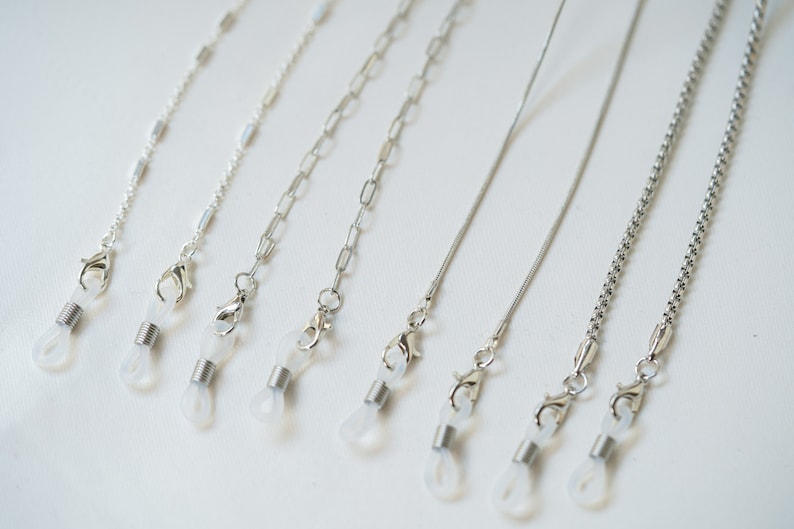 May include: Seven silver eyeglass chains with clear plastic clasps. The chains are different styles, including a thin chain, a thicker chain, and a chain with small links.