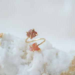May include: A gold ring with two pink, raw, irregular shaped stones set in the band. The ring is on a white and brown rock.