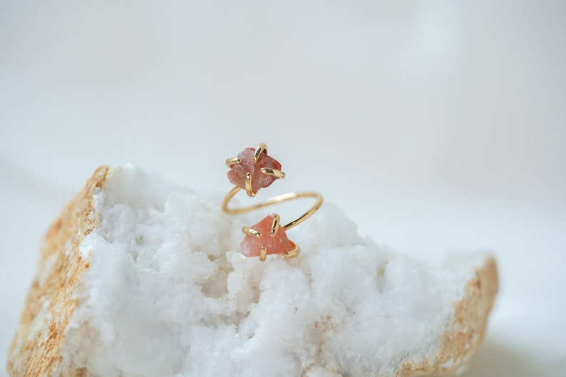May include: A gold ring with two pink, raw, irregular shaped stones set in the band. The ring is on a white and brown rock.