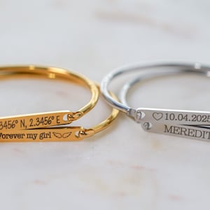 May include: Two personalized bangles, one gold-toned and one silver-toned, with engraved text. The gold bangle reads "12.3456° N, 2.3456° E" and "Forever my girl". The silver bangle reads "10.04.2025" and "MEREDITH".