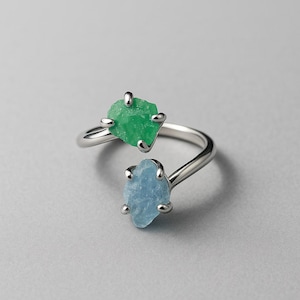 May include: A silver ring featuring two raw gemstones: a vibrant green and a light blue. The ring's band curves around the finger, with each stone held in place by four small prongs. The gemstones have a rough, natural texture.