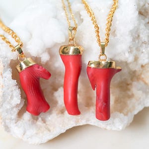 May include: Four gold necklaces with red coral pendants. The coral is shaped like a branch and has a natural, organic look. The necklaces are displayed on a white crystal background.