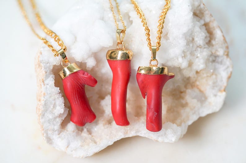 May include: Four gold necklaces with red coral pendants. The coral is shaped like a branch and has a natural, organic look. The necklaces are displayed on a white crystal background.