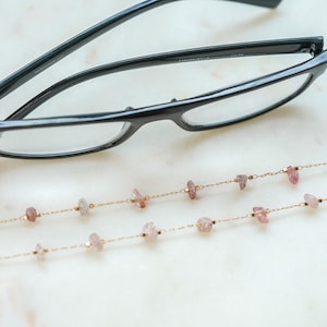 May include: A pair of black eyeglasses with a gold chain featuring pink and white gemstone beads. The chain is designed to hold the eyeglasses around the neck.