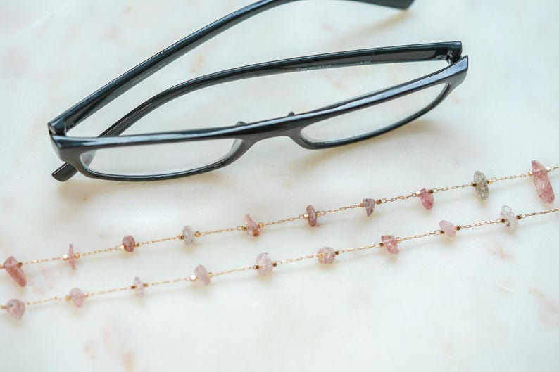 May include: A pair of black eyeglasses with a gold chain featuring pink and white gemstone beads. The chain is designed to hold the eyeglasses around the neck.