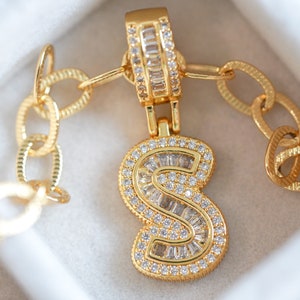 May include: Gold chain necklace with a pendant in the shape of the letter "S". The pendant is encrusted with small, clear cubic zirconia stones.