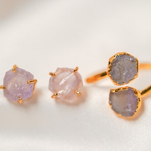 May include: A set of three gold-toned rings featuring raw amethyst stones. The rings are displayed on a white fabric background.
