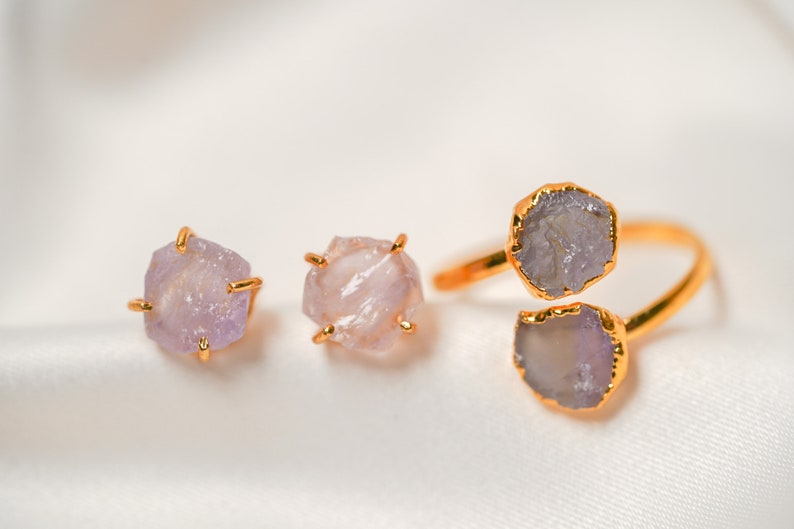May include: A set of three gold-toned rings featuring raw amethyst stones. The rings are displayed on a white fabric background.