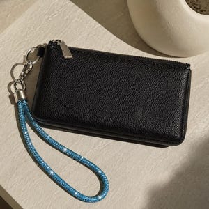 May include: A rectangular black pebbled leather wallet featuring a silver zipper closure and a detachable wrist strap. The wrist strap is covered in small, sparkling blue crystals and is attached to the wallet with a silver metal ring and clasp.