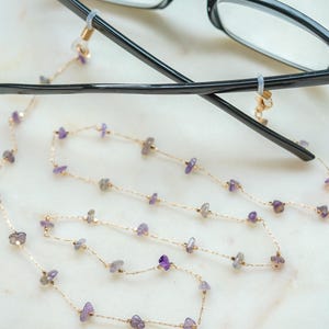 May include: A gold chain with small purple gemstones, attached to a pair of black eyeglasses with clear lenses. The chain is designed to hold the glasses around the neck.