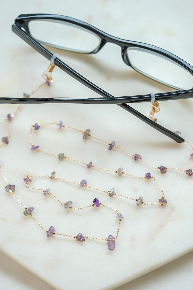 May include: A gold chain with small purple gemstones, attached to a pair of black eyeglasses with clear lenses. The chain is designed to hold the glasses around the neck.