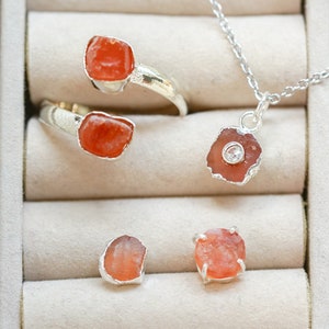 May include: A set of four silver rings with orange gemstone accents. The rings are displayed in a white box with velvet lining. One ring is a double band with two orange stones. The other three rings each feature a single orange stone.