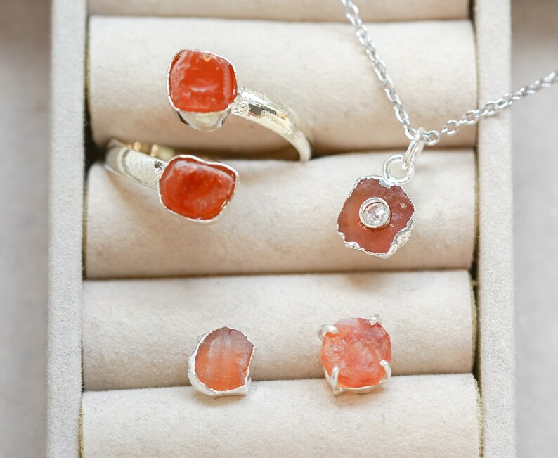 May include: A set of four silver rings with orange gemstone accents. The rings are displayed in a white box with velvet lining. One ring is a double band with two orange stones. The other three rings each feature a single orange stone.