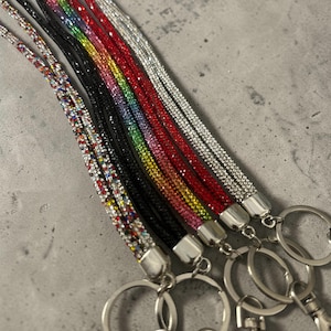 May include: A selection of rhinestone lanyards in various colours, including silver, black, red, pink, yellow, green, and a rainbow design. Each lanyard has a silver-toned metal clasp and a key ring. The lanyards are for holding keys or ID badges.