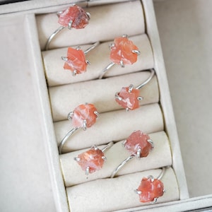 May include: A set of eight silver rings with raw, orange-red gemstone settings. The rings are displayed in a white box with individual compartments.