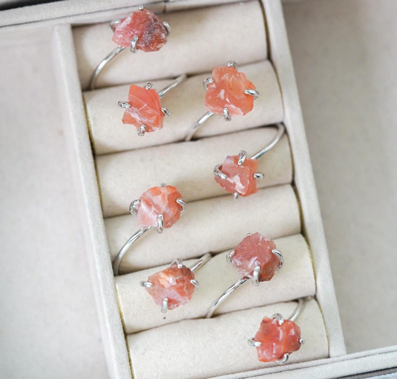 May include: A set of eight silver rings with raw, orange-red gemstone settings. The rings are displayed in a white box with individual compartments.