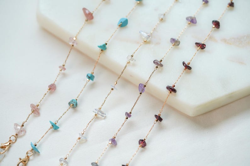 May include: Four gold chains with different colored gemstone beads. The chains are laid out on a white marble surface.
