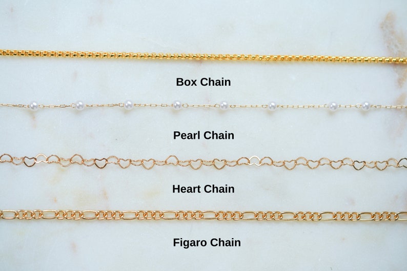 May include: Four different types of gold chains are displayed on a white marble surface. The chains are labeled "Box Chain", "Pearl Chain", "Heart Chain", and "Figaro Chain".