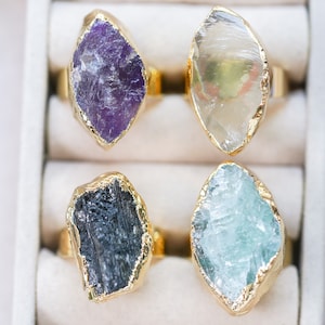 Op de afbeelding: Vier edelsteenringen met goudkleurige banden. De edelstenen hebben verschillende kleuren, waaronder paars, helder, zwart en blauw. De ringen worden gepresenteerd in een witte sieradendoos, die de unieke texturen en kleuren van de stenen benadrukt.