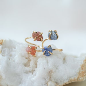 May include: Three gold rings with different colored stones. The rings are stacked on top of each other. The stones are a light pink, a dark blue, and a light blue.
