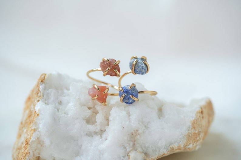 May include: Three gold rings with different colored stones. The rings are stacked on top of each other. The stones are a light pink, a dark blue, and a light blue.