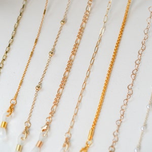 May include: A set of nine gold-tone eyeglass chains with different styles, including a chain with small beads, a chain with heart-shaped links, and a chain with small round beads. The chains are all about 25 inches long and have clear plastic clasps.