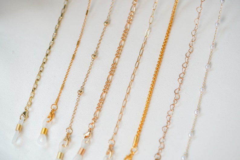 May include: A set of nine gold-tone eyeglass chains with different styles, including a chain with small beads, a chain with heart-shaped links, and a chain with small round beads. The chains are all about 25 inches long and have clear plastic clasps.