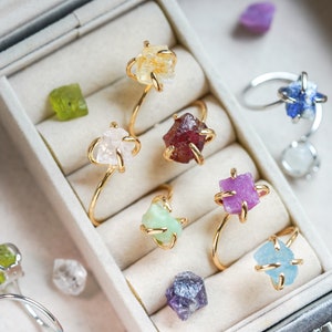 May include: A collection of gemstone rings in a jewelry box. The rings feature raw, uncut gemstones in various colors, including yellow, pink, red, green, purple, and blue. The rings have gold or silver bands. Scattered gemstones of different colors are also visible.