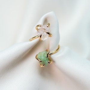 May include: Two gold-colored rings with raw gemstone settings. One ring features a light pink stone, and the other has a green stone. The rings are set against a white, textured background, showcasing the jewelry's natural aesthetic.