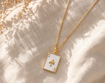 Puede incluir: Un collar de cadena tipo caja en tono dorado con un colgante rectangular en forma de carta de juego. El colgante tiene una cara de esmalte blanco con un símbolo de as de picas en tono dorado en el centro y pequeñas marcas en las esquinas, suspendido de una cadena fina.