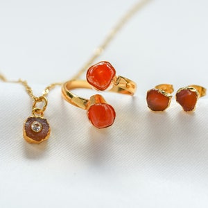 May include: A set of gold jewelry featuring three pieces with orange carnelian stones. The set includes a ring with two stones, a pair of earrings, and a pendant necklace.